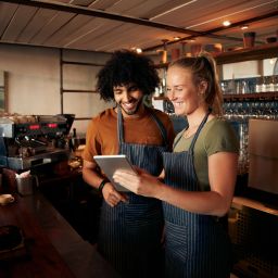 Smiling waiter and waitress wearing apron using digital tablet standing behind counter in cafe