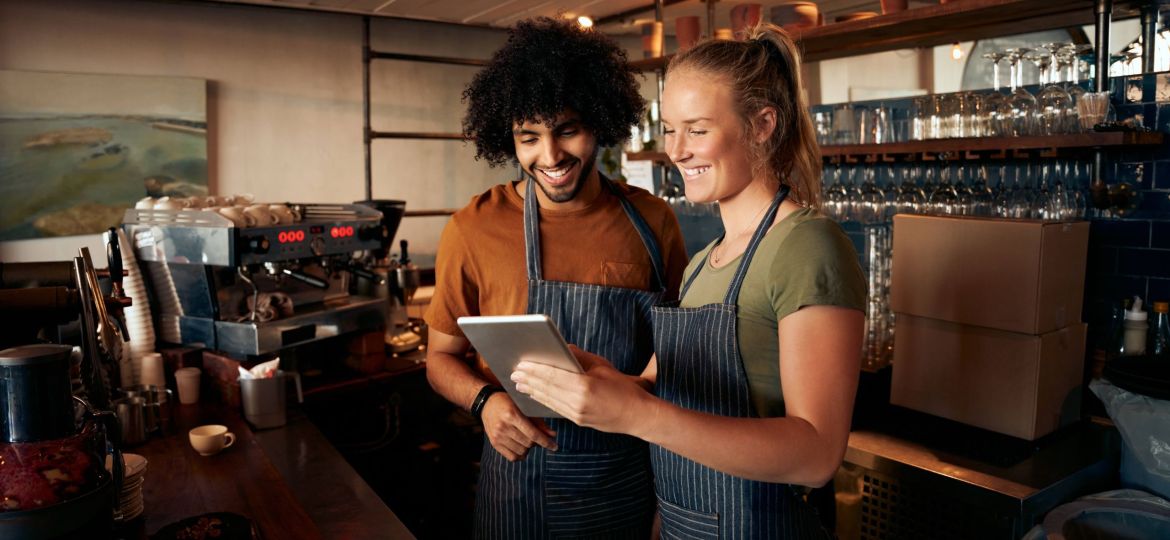 Smiling waiter and waitress wearing apron using digital tablet standing behind counter in cafe