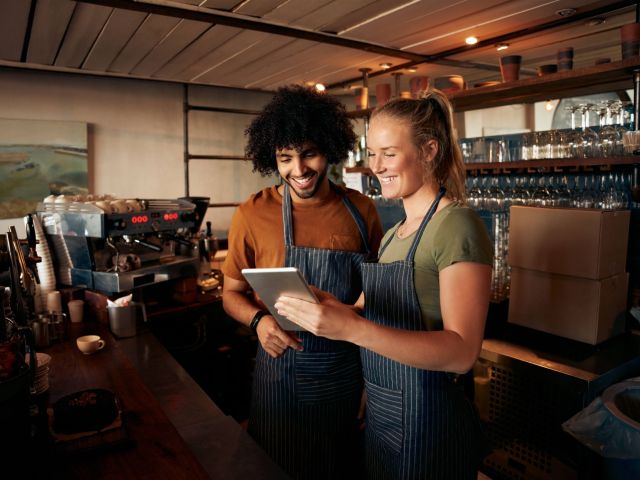 Smiling waiter and waitress wearing apron using digital tablet standing behind counter in cafe