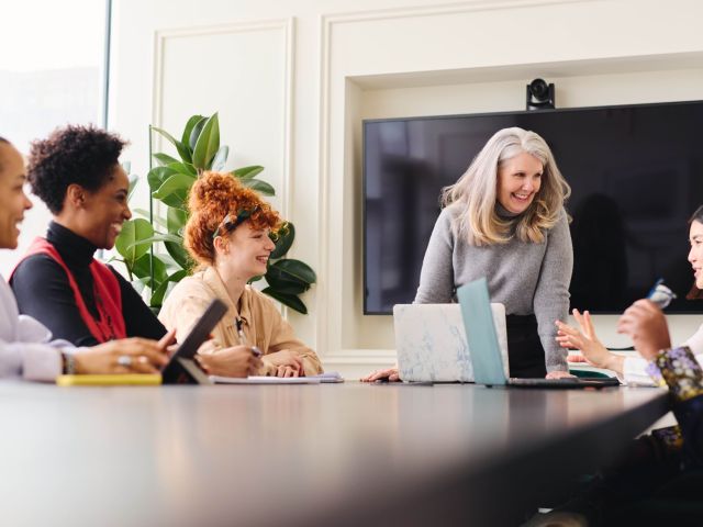 Portrait of senior white businesswoman with laptop smiling and listening to multiethnic female colleagues in boardroom